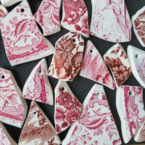 Può includere: Una collezione di frammenti di ceramica bianca con motivi floreali e botanici rossi. Ogni frammento ha un foro per essere appeso. I motivi includono fiori, foglie e altri elementi decorativi. I frammenti sono disposti su una superficie scura.