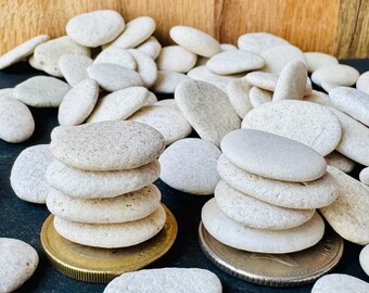 May include: A collection of smooth, light-colored river stones. Several stacks of stones are balanced on coins, with more stones scattered around. The background is a wooden surface.