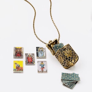 May include: A gold-toned metal locket pendant on a chain, designed as a miniature hinged box with an intricate floral filigree pattern. Inside the locket and scattered around are tiny rectangular tarot cards featuring illustrations and text labels.
