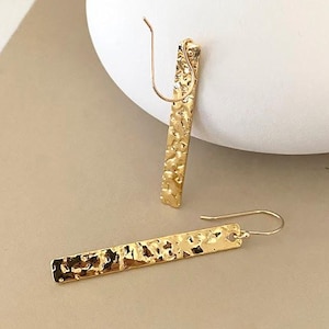 May include: A pair of gold-colored, hammered-metal earrings. Each earring is a long, rectangular bar with a textured surface and a curved ear wire. The earrings are displayed on a neutral background.