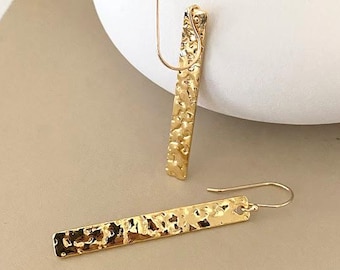 May include: A pair of gold-colored, hammered-metal earrings. Each earring is a long, rectangular bar with a textured surface and a curved ear wire. The earrings are displayed on a neutral background.