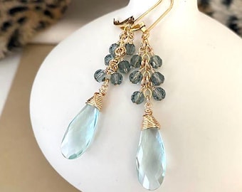 May include: Gold-tone earrings with a cluster of small, gray beads and a large, teardrop-shaped, light blue crystal. The crystals are wrapped in gold wire. The earrings are displayed on a white ceramic vase.