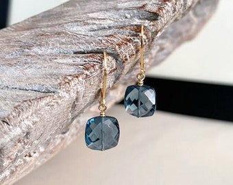 Puede incluir: Un par de pendientes de oro con gotas de piedras preciosas azules. Las piedras preciosas son facetadas y tienen forma cuadrada.