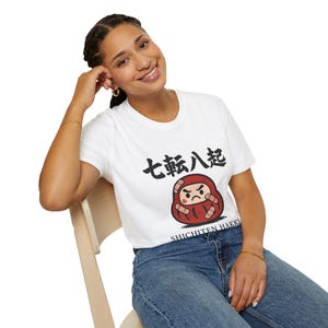 May include: White t-shirt featuring a Japanese Daruma doll design with the text "Shichiten Hakki". The Daruma doll is red with a grumpy expression. The shirt is made of a soft material.
