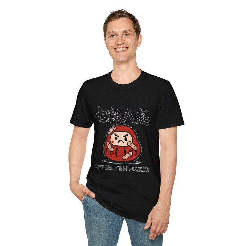 May include: Black t-shirt featuring a red Daruma doll illustration with the Japanese text "Shichiten Hakki" and other Japanese characters. The Daruma doll has a stern expression and is surrounded by bandages.