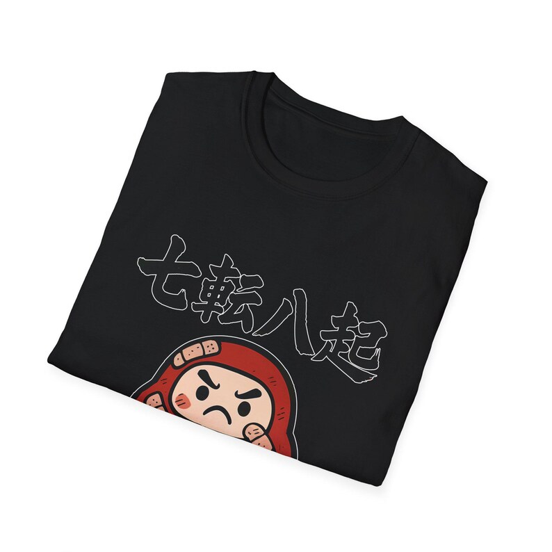 May include: Black t-shirt featuring a Japanese Daruma doll design with the text "七転八起" in white. The Daruma doll is red with a grumpy expression. The shirt is folded, showing the design on the front.