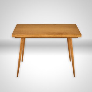 Op de afbeelding: Een rechthoekige houten tafel met een gladde, lichtbruine afwerking. De tafel heeft een vlak bovenblad en vier slanke, taps toelopende poten die vanuit het midden naar buiten staan, wat zorgt voor een stabiele basis.