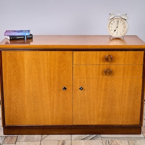 Può includere: Un mobile in legno marrone chiaro con due ante e due cassetti. Una sveglia bianca e due libri sono sopra. Il mobile ha una finitura e una base marrone scuro. L'orologio segna le 7:07.