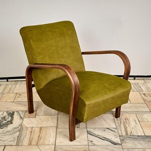 Op de afbeelding: Olijfgroene fauteuil met gebogen houten armleuningen en poten. De stoel heeft een afgeronde rugleuning en een gewatteerde zitting. Het houten frame heeft een warme, donkere afwerking die contrasteert met de groene stof.