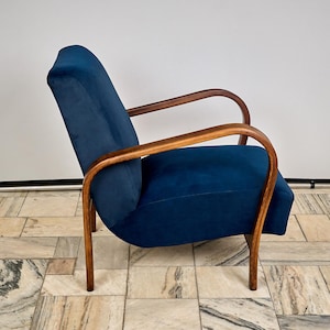 Op de afbeelding: Een vintage fauteuil met een donkerblauwe fluwelen zitting en rugleuning. De stoel heeft gebogen, lichtbruine houten armleuningen en poten. Het ontwerp is strak en minimalistisch, geschikt voor een woonkamer of lounge. Een klassieker uit het midden van de eeuw.