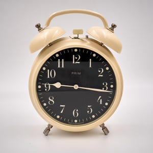 May include: Cream-colored vintage alarm clock with a black face and white numerals. The clock has a curved handle, two bells, and a base with small legs. The word "PRIM" is printed on the clock face.