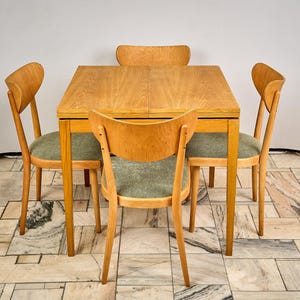 Puede incluir: Una mesa de comedor de madera clara con cuatro sillas a juego. La mesa es cuadrada con un acabado de veta de madera clara. Las sillas tienen respaldos curvos y asientos tapizados verdes. El conjunto está sobre un suelo de baldosas estampadas. La mesa mide unos 76 cm de lado.