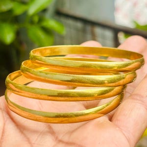 May include: A set of three smooth, polished gold-tone bangles. Each bangle features a simple, rounded, circular design with a reflective metallic finish. The bracelets are stacked together, showcasing a consistent yellow gold color throughout.