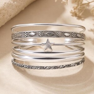 May include: A silver-toned cuff bracelet featuring multiple stacked bands. The design includes two textured bands with intricate floral and leaf patterns, three smooth polished bands, and a central band adorned with a single five-pointed star.