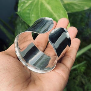Puede incluir: Un brazalete de plata pulida con un diseño curvo y abierto. El brazalete tiene una superficie lisa y reflectante y se sostiene en una mano sobre un fondo de follaje verde. El brazalete mide aproximadamente 6,35 cm de ancho.
