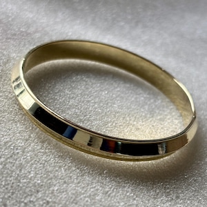 May include: A gold-colored bangle bracelet. The bracelet is a solid band with a smooth, reflective surface. The bangle is approximately 2.5 inches in diameter and has a simple, elegant design. The bracelet is likely made of metal.