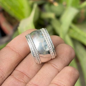 May include: A wide, silver ring with a textured, braided design on the edges. The ring has a smooth, polished centre band. The ring is held in a hand, with a blurred green background.