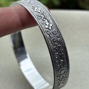 May include: A silver-colored bangle bracelet with an intricate floral design. The bracelet features detailed engravings of flowers and vines, creating a textured surface. The edges have a beaded border, adding to the ornate style.