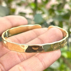 Könnte beinhalten: Ein goldfarbenes Armreif-Armband mit gehammerter Textur. Das Armband hat ein schlichtes, offenes Design und wird auf einer Hand gezeigt. Das Metall reflektiert das Licht und erzeugt Glanzlichter und Schatten.