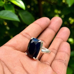 Puede incluir: Un anillo de plata con una gran piedra preciosa azul oscuro rectangular y facetada, sujeta por cuatro garras de metal. La piedra tiene una superficie pulida con bordes geométricos definidos y el aro es de acabado metálico plateado liso.