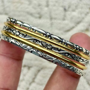May include: A collection of silver and gold bangles. The silver bangles feature a floral and leaf pattern. The gold bangles are plain. The bangles are stacked, creating a layered effect. The jewellery is held by a person's fingers.