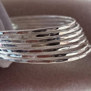 Puede incluir: Una pila de brazaletes plateados con una textura martillada. Los brazaletes son redondos y tienen una superficie ligeramente irregular, que refleja la luz. La pila es parcialmente visible, mostrando el diseño en capas y el acabado metálico pulido.