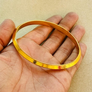 Peut inclure: Un bracelet jonc de couleur or avec un profil plat et rectangulaire. Le bracelet est une bande d'or massif, reflétant la lumière. Le jonc est tenu dans une main, mettant en valeur son design simple et élégant. Ce bijou est adapté au quotidien.