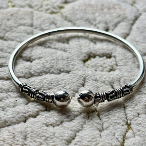 Op de afbeelding: Een zilverkleurige open manchetarmband. De armband heeft decoratieve details aan de uiteinden, met een afgeronde bal aan elke opening. De armband is ontworpen om om de pols te worden gedragen.