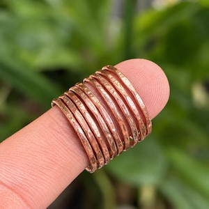 Peut inclure: Un ensemble de dix bagues en cuivre individuelles, chacune présentant une texture martelée distincte. Les bagues sont de fines bandes circulaires avec une finition métallique brun rougeâtre, disposées côte à côte en formation empilée.