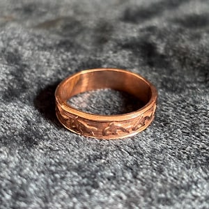 May include: A copper-coloured ring with an engraved mountain range design. The ring is circular and sits on a dark grey, textured surface. The ring's band features a detailed relief of peaks and valleys.