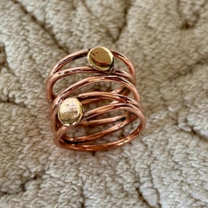 May include: A stack of copper rings with two gold-coloured oval accents. The rings have a textured finish and are arranged in a spiral. The jewellery is displayed on a textured, neutral-coloured surface.