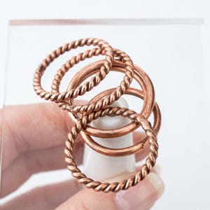 Op de afbeelding: Een set van drie koperkleurige ringen die op elkaar zijn gestapeld. Twee van de ringen hebben een gedraaide, touwachtige textuur, terwijl de derde ring een glad, effen oppervlak heeft. De ringen zijn van metaal met een warme, roodbruine afwerking.