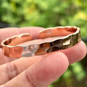 May include: A hammered copper cuff bracelet with a textured surface. The open cuff design features irregular shapes and a polished finish. The bracelet is held in a hand against a blurred green background.