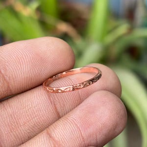May include: A rose gold ring with a textured, patterned design. The ring is thin and delicate, with a repeating pattern around the band. The ring is held in a hand, with a blurred green background.