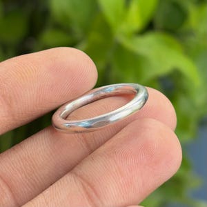 Puede incluir: Un anillo de plata con un diseño liso y redondeado. El anillo se sostiene entre los dedos, con un fondo verde borroso. La superficie del anillo refleja la luz, resaltando su acabado pulido. Una joya sencilla y elegante.