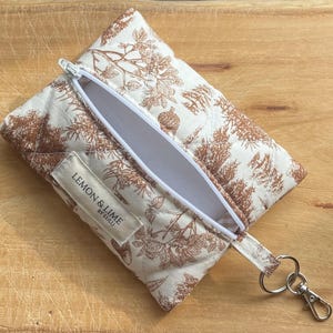 May include: A quilted rectangular pouch featuring a brown botanical print of acorns and leaves on a cream background. The pouch has a white zip, a white interior lining, and a fabric label reading LEMON & LIME BY LULU. A metal keyring is attached.