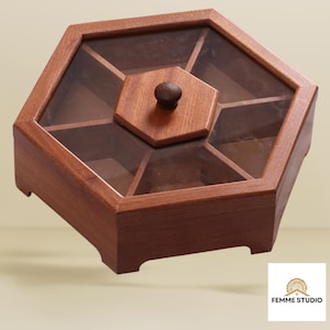 May include: A wooden, hexagonal serving tray with a clear glass top and a small, round knob. The tray is divided into six sections and is made of a dark brown wood. The Femme Studio logo is in the bottom right corner.