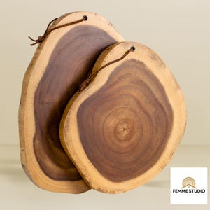 Op de afbeelding: Twee houten snijplanken met natuurlijke randen. De planken zijn gemaakt van donkerbruin hout met lichtere bruine randen. Elke plank heeft een leren lus om op te hangen. De planken hebben verschillende afmetingen.