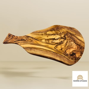 May include: Olive wood serving board with a natural, organic shape. The board features a handle and a rich, swirling grain pattern in shades of brown and gold. The board is ideal for serving appetizers or charcuterie.