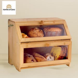 May include: A two-tiered bamboo bread box with clear glass windows. The top section holds a croissant and two loaves of bread. The bottom section contains sliced bread and a baguette. The box has a wooden knob and the text "FEMME STUDIO".