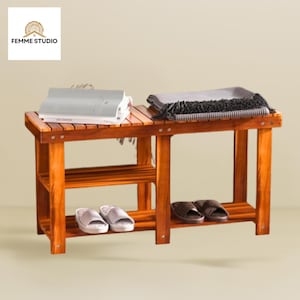 May include: A wooden bench with two shelves, holding a pair of white and a pair of brown slippers. A gray blanket and a book rest on the bench's top. The bench has a warm, reddish-brown finish. The logo "FEMME STUDIO" is in the top left corner.