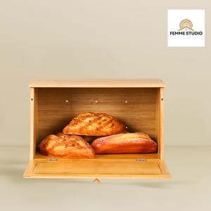 May include: A wooden bread box with three loaves of bread inside. The bread box is open, revealing the golden-brown crusts of the bread. The box is made of light-colored wood and has a simple, rectangular design.