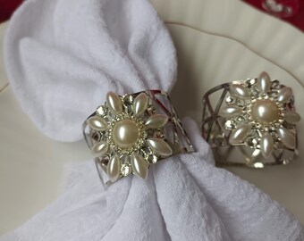 Könnte beinhalten: Zwei silberne Serviettenringe mit Perlen- und Kristallverzierungen schmücken eine weiße Leinen-Serviette auf einem weißen Teller. Die Serviettenringe haben ein Blumenmuster mit einer großen Perle in der Mitte. Der Hintergrund ist einfarbig rot.