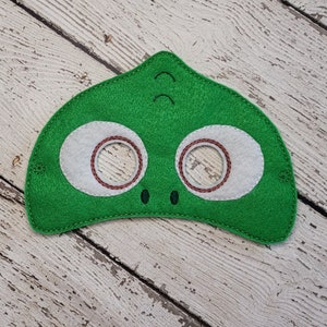 May include: A green felt mask featuring two large white eye patches with brown stitched circular outlines, two small dark green stitched nostrils, and two small dark green stitched curved lines above the eyes, with side holes for attachment.