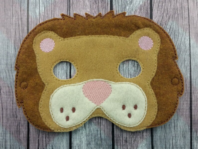 May include: A felt lion mask featuring a dark brown mane, a tan face, and a cream-coloured muzzle. The mask includes two eye cutouts, pink embroidered ears, a pink embroidered nose, and small brown stitched dots representing whiskers on the muzzle.
