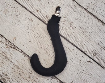 Op de afbeelding: Een zwart met stof bekleed accessoire in de vorm van een J-haak, voorzien van een zilverkleurige metalen clip aan de bovenzijde. Het item heeft een gladde, matte textuur en een gebogen, afgerond ontwerp voor functionele bevestiging.