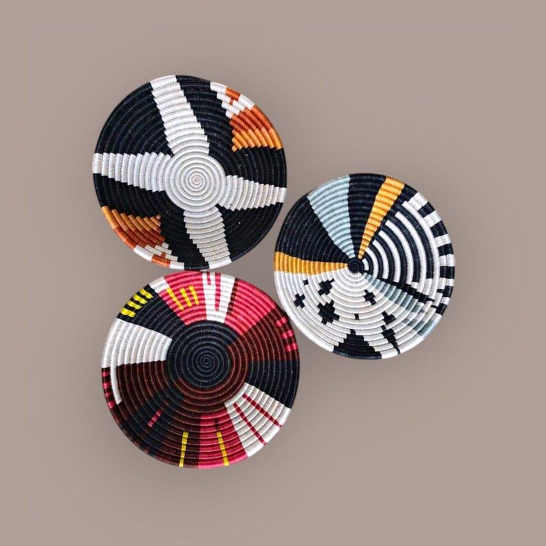 May include: Three round woven baskets with geometric patterns. One basket features black, white, and orange segments. Another has black, white, yellow, and blue sections. The third basket displays red, black, white, and yellow designs.