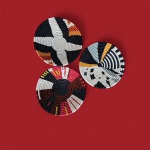 May include: Three decorative woven baskets with geometric patterns in black, white, red, and orange. The baskets are circular and arranged on a solid red background. The designs include stripes, stars, and radial patterns.