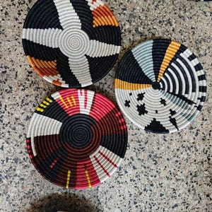 May include: A collection of woven, circular coasters with geometric patterns. The coasters feature a variety of colors, including black, white, red, orange, and yellow. The coasters are made of a natural fiber material.