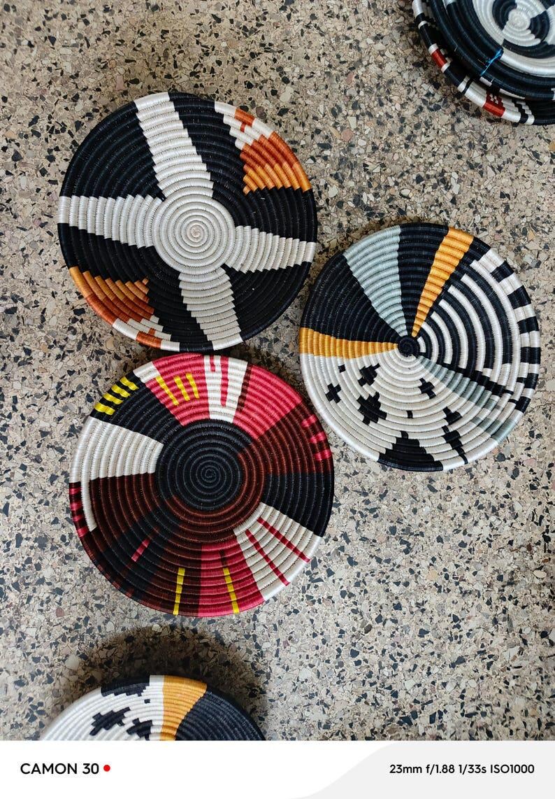 May include: A collection of woven, circular coasters with geometric patterns. The coasters feature a variety of colors, including black, white, red, orange, and yellow. The coasters are made of a natural fiber material.